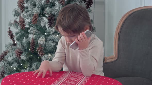 Cute Little Boy Calling Santa While Sitting on a Big Armchair at Home Over Chirstmas Tree Background