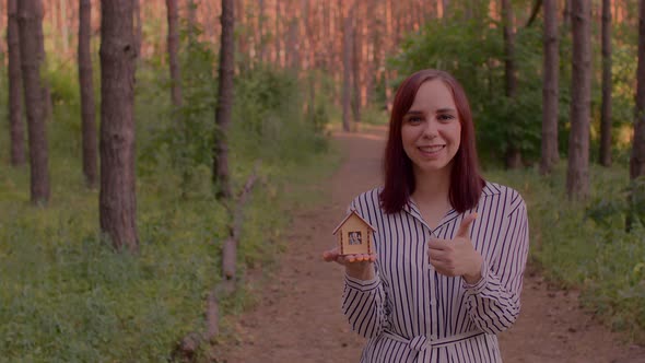 Young Woman Holding Small Wooden House and Showing Thumb Up in Forest