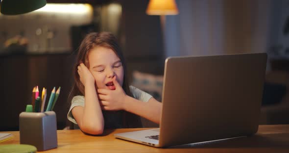 Preteen Girl Yawning Doing Homework at Table with Laptop in Evening