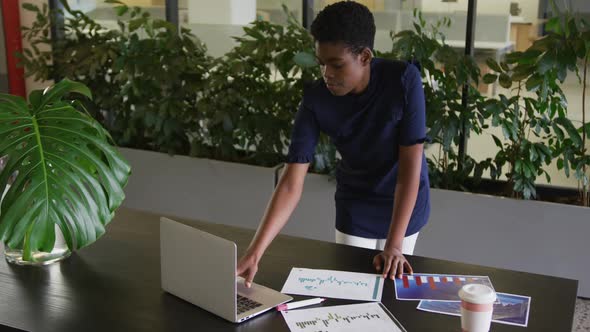 African american businesswoman standing using laptop going through paperwork in modern office