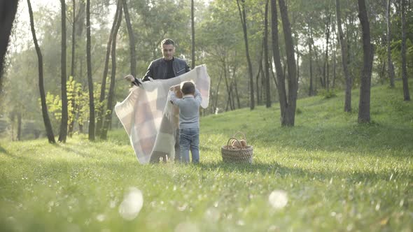 Wide Shot of Caucasian Young Father and Little Son Laying Blanket on Green Summer Meadow