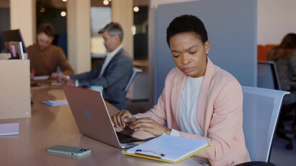 African American Concentrated Woman Looking at Clipboard Working on the Laptop While Sitting at