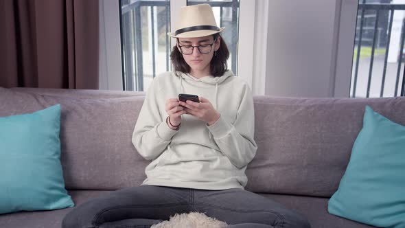 Brunette Girl in a Hat Uses Her Cell Phone at Home on the Couch Window Background