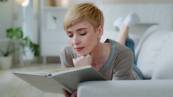 Serene Young Woman Relaxing at Home Alone Lying on Sofa Holding in Hands Open Book Reading New