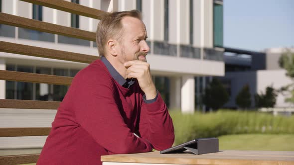 A Middleaged Handsome Caucasian Man Looks at a Tablet and Thinks As He Sits at a Table in a Park