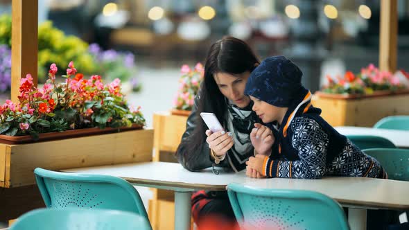 A Young Mother with Smart Phone Showing Something Her Little Cute Son at a Table in a Cafe