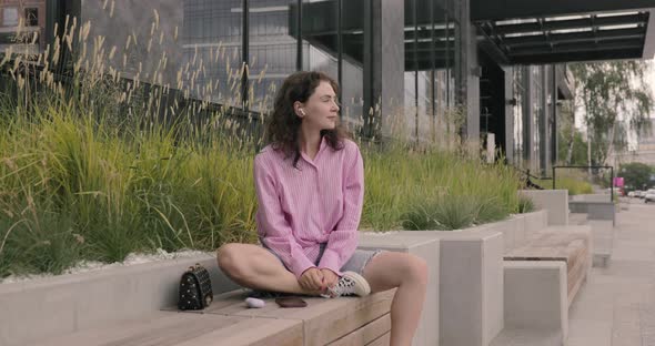 Portrait of joyful woman wearing pink shirt  listening music, sit at the bus stop.