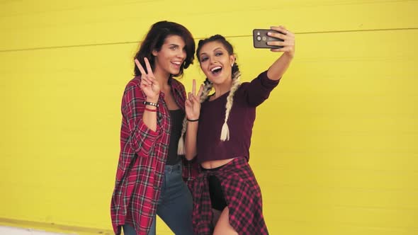 Two Hipster Girls Making Selfie and Showing Peace Sign Standing By the Yellow Wall