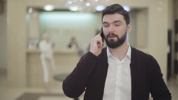 Handsome Caucasian Brunette Man with Beard Hanging Up Phone and Smiling at Camera at the Background