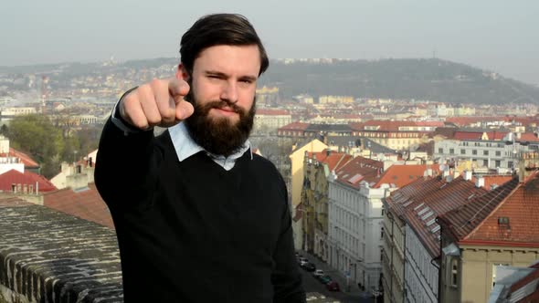 Young Handsome Man with Full-beard (Hipster) Points To Camera - City in Background