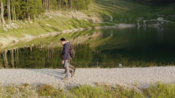 Couple Hiking Past Lake On Mountain Petzen Surrounded By Trees. Aerial Follow Shot