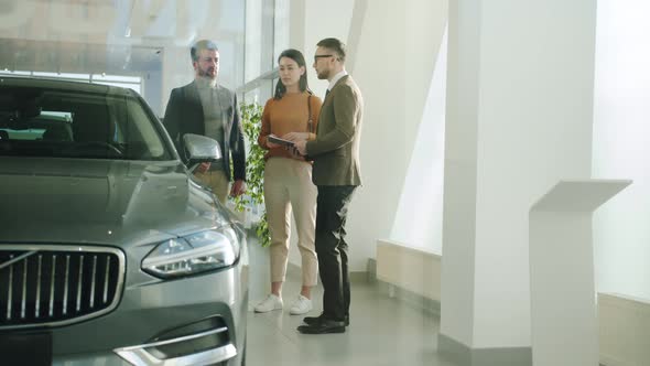 Girl and Guy Talking To Salesman in Car Dealership Choosing Automobile Together