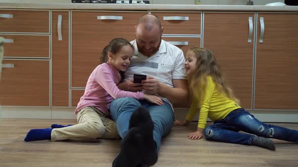 the Young Father and His Happy Daughters Look at Their Smartphones Sitting in the Kitchen