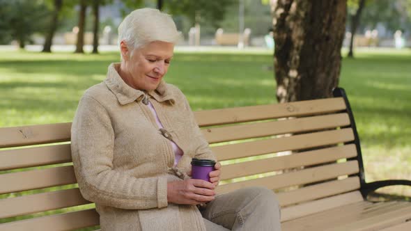 Dreamy Calm Relaxed Grayhaired Elderly Grandmother Sitting on Park Bench Drinking Coffee From Glass