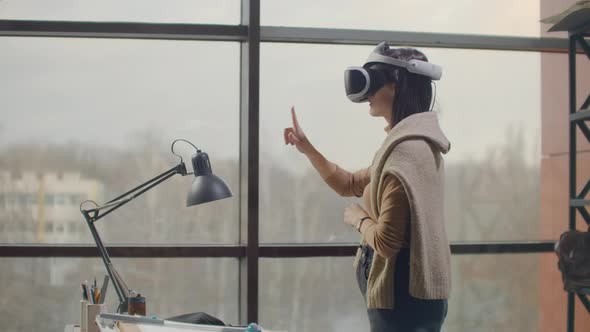 Female Engineer in a Virtual Reality Helmet Near the Large Window with His Hands Imitates the Work