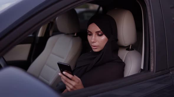 Attractive Young Woman in Black Hijab Sitting in Her Car Thoughtfully Looking at Her Smartphone