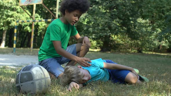 African Boy Comforting Teenage Friend Suffering Pain Lying Ground, Sport Injury