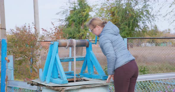 Young Blonde Woman in Down Jacket Opens a Well and Lowers Zinc Bucket on a Rope Using Special Manual