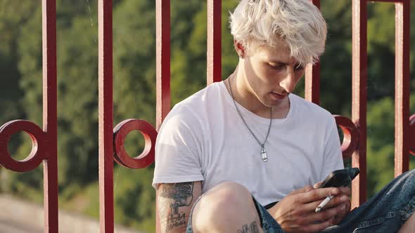 Young Fashionable Blonde with Tattoos Smokes a Cigarette and Holds a Smartphone in His Hands While