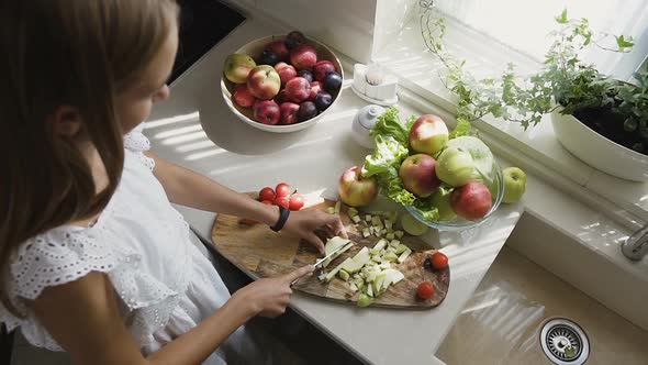Attractive Blonde Girl in White Dress is Preparing Fruits Salad in the Kitchen