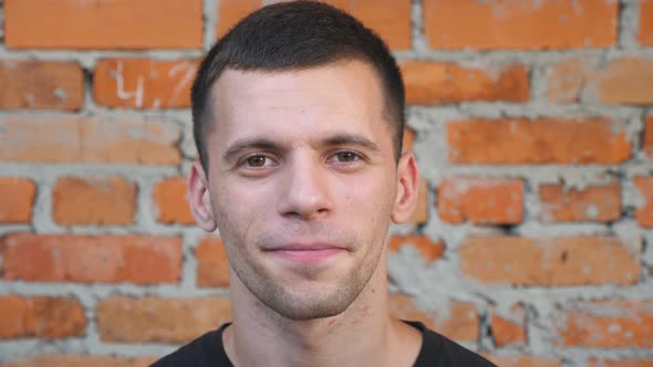 Portrait of Smiles Young Man with Brown Hair Standing on Blurred Brick Wall Background. Close Up of