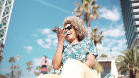 Happy African grey haired woman wearing sunglasses recording voice message by mobile