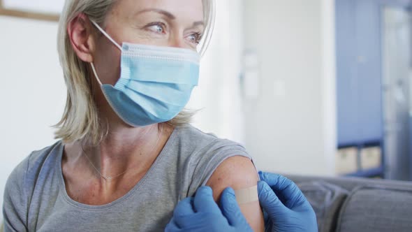 Happy senior diverse woman and doctor wearing face masks in living room sitting on sofa, vaccinating