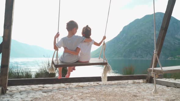 A Girl and a Boy Swing on a Swing Near the Sea with a Beautiful View of the Bay and Mountains