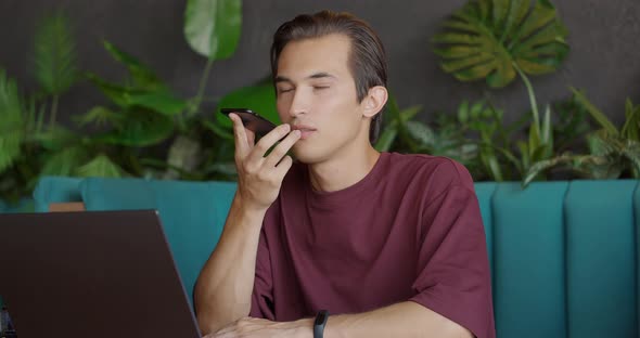 Handsome Young Man in Eyeglasses is Sitting at a Table in a Cafe and Talking in a Voice Chat Via