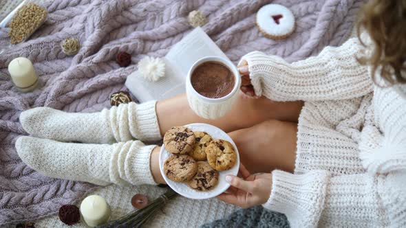 Woman's Hands And Feet In Sweater And Knit Cozy Beige Socks Holding Cup Of Hot Chocolate And Cookies