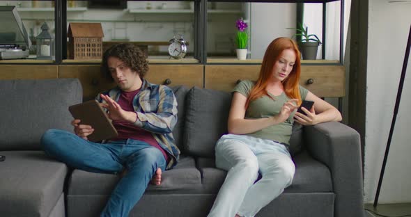 Young Man and Woman Are Sitting on Sofa Edges, Using Their Gadgets