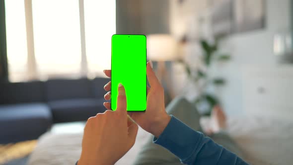 Woman at Home Lying on a Bed and Using Smartphone with Green Mockup Screen in Vertical Mode