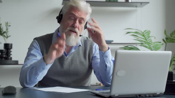 Smiling Elderly Man with a Gray Beard Wearing Headphones, Having a Video Call on a Laptop, Happy
