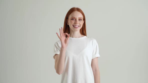 Portrait of Smiling Redhead Young Woman Showing Okay Gesture Looking at Camera Standing on White