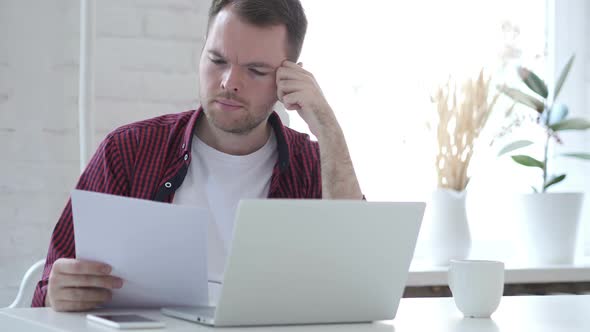 Young Man Reading Documents and Working on Laptop