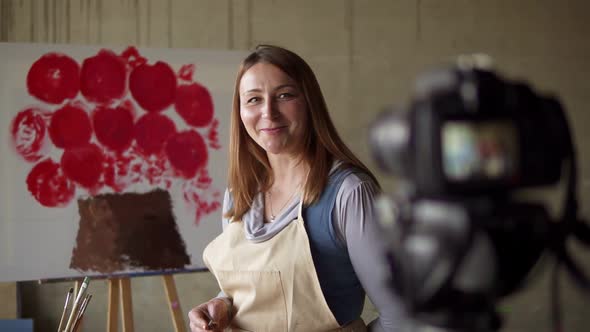 Female Caucasian Mature Blogger Recording Video in Workshop Drawing Pink Flower on Canvas Explaining