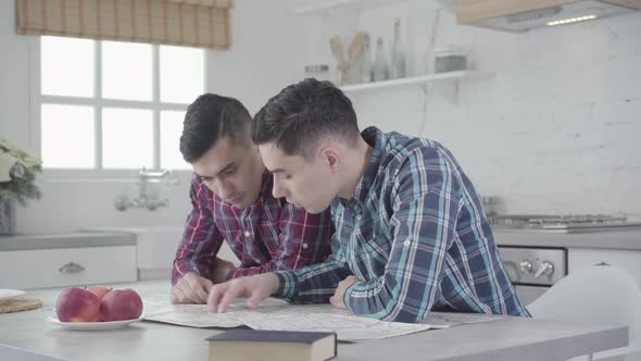 Portrait of Two Caucasian Twin Brothers Sitting at the Table and Looking at Map. Siblings Planning