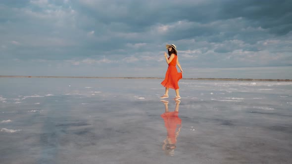 Young Woman Tourist in a Waving Dress Walks Along a Salt Lake in Which the Sky Is Reflected