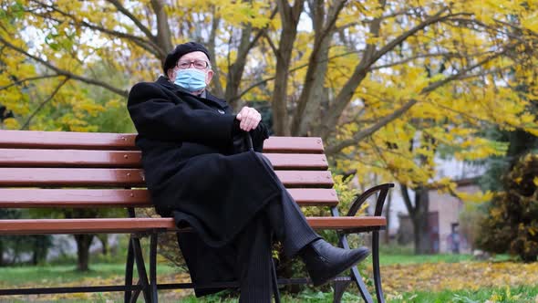 An Old Grandfather in a Black Coat and Beret He is Sitting on a Park Bench in a Protective Mask