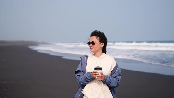 Beautiful Young Woman in the Morning Stands on the Beach with Black Volcanic Sand and Drinks Coffee