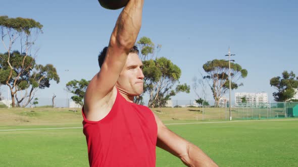 Fit caucasian man exercising outdoors, squatting and lifting kettlebell weight with one arm