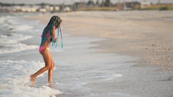 A Girl with African Braids in a Summer Costume Plays on the Beach with Shells Near the Sea with