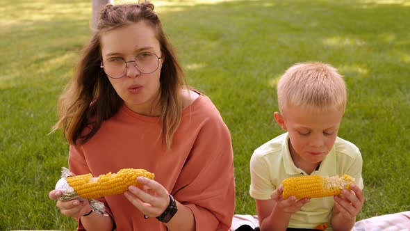 A Teenage Girl and a Little Boy are Eating Corn in a Park Sitting on the Grass