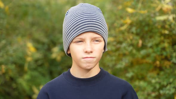 Serious Boy in Striped Hat Standing in Park Looks at Camera and Nods His Head in Agreement