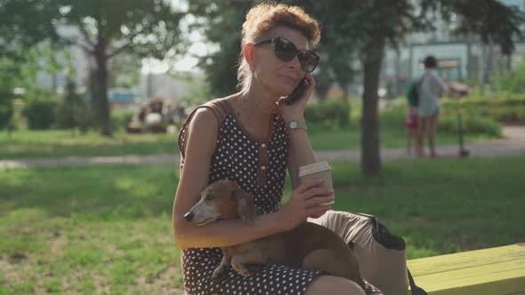 Elderly Woman Drinking Coffee to Herself Sitting on a Bench Holding a Dachshund Dog in the Park in