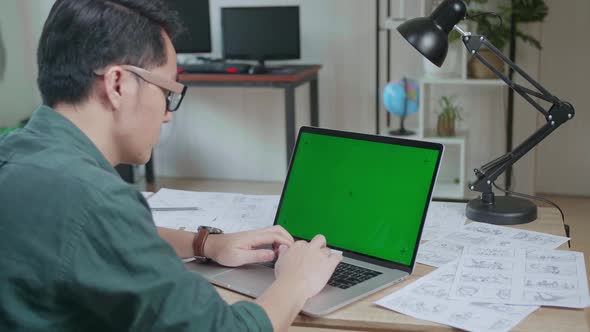 Asian Man Works On Laptop Computer With Mock Up Green Screen Display And Storyboard