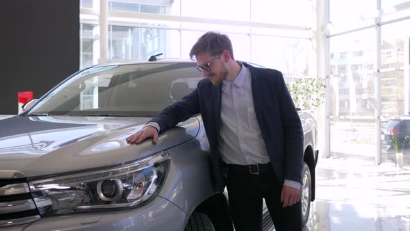 Auto Purchase, Young Buyer Man with Glasses Gladly Strokes New Car and Gives Positive Gesture