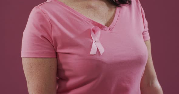 Mid section of a woman with pink ribbon on her chest clenching her fist against pink background