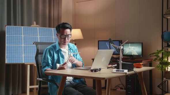 Asian Man Sitting In Front Of Solar Cell Look At Wind Turbine While Typing On A Laptop At The Office