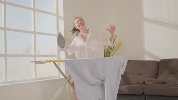Cheerful woman in man's shirt dancing when ironing clothes in room at home.